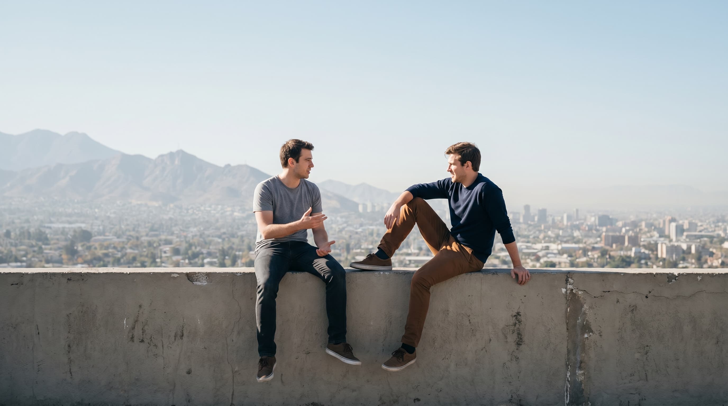 Two men sitting on a rooftop wall in casual conversation with city skyline and mountains in the background, representing BARGOBET's urban lifestyle for tall men